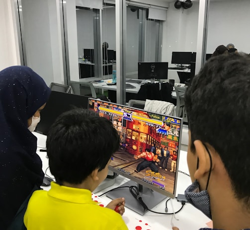 A group of children is gathered around a desktop computer, engaged in playing a colorful, action-packed arcade game displayed on the monitor. The setting appears to be a modern, well-lit office or gaming space with large glass windows, showing multiple computer workstations in the background.